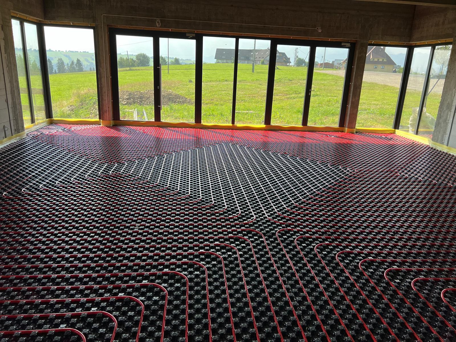 Czerwone rury ogrzewania podłogowego ułożone na czarnej macie w surowym wnętrzu z widokiem na zielone wzgórza przez duże okna.