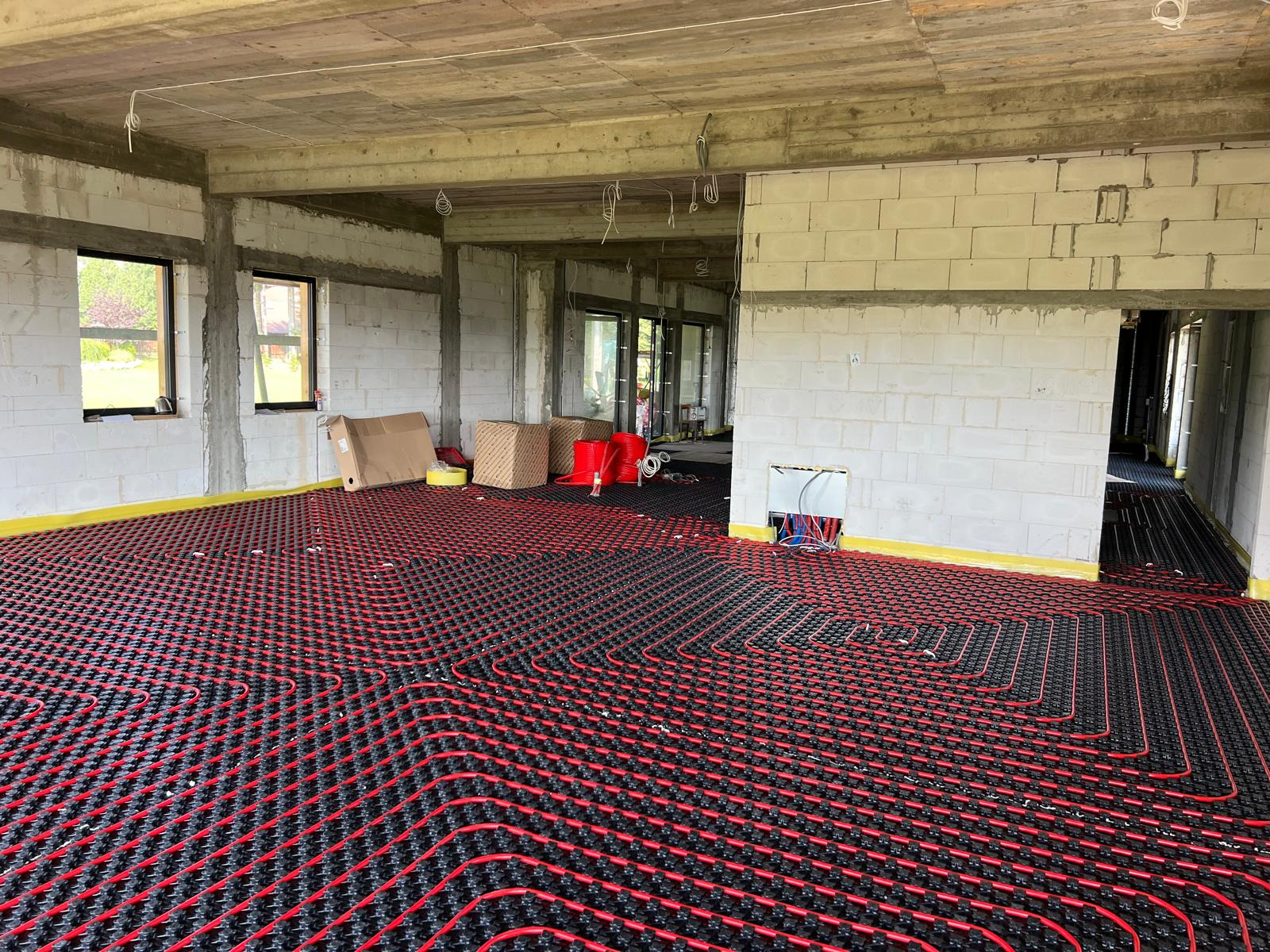 Czerwono-czarna instalacja ogrzewania podłogowego w surowym wnętrzu z białych bloczków, widoczne okna i kable. Żółta taśma wzdłuż ścian.