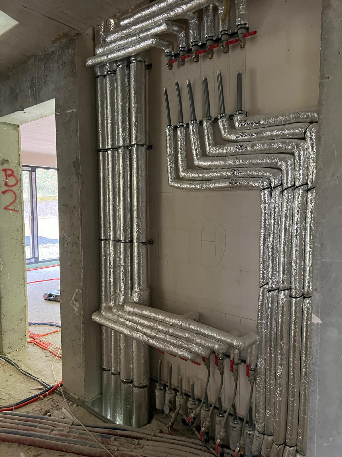 Izolowane rury instalacji grzewczej, prowadzone wzdłuż surowej ściany, z widocznymi zaworami i manometrami. Instalacja w trakcie montażu, widoczne oznaczenia na ścianie.