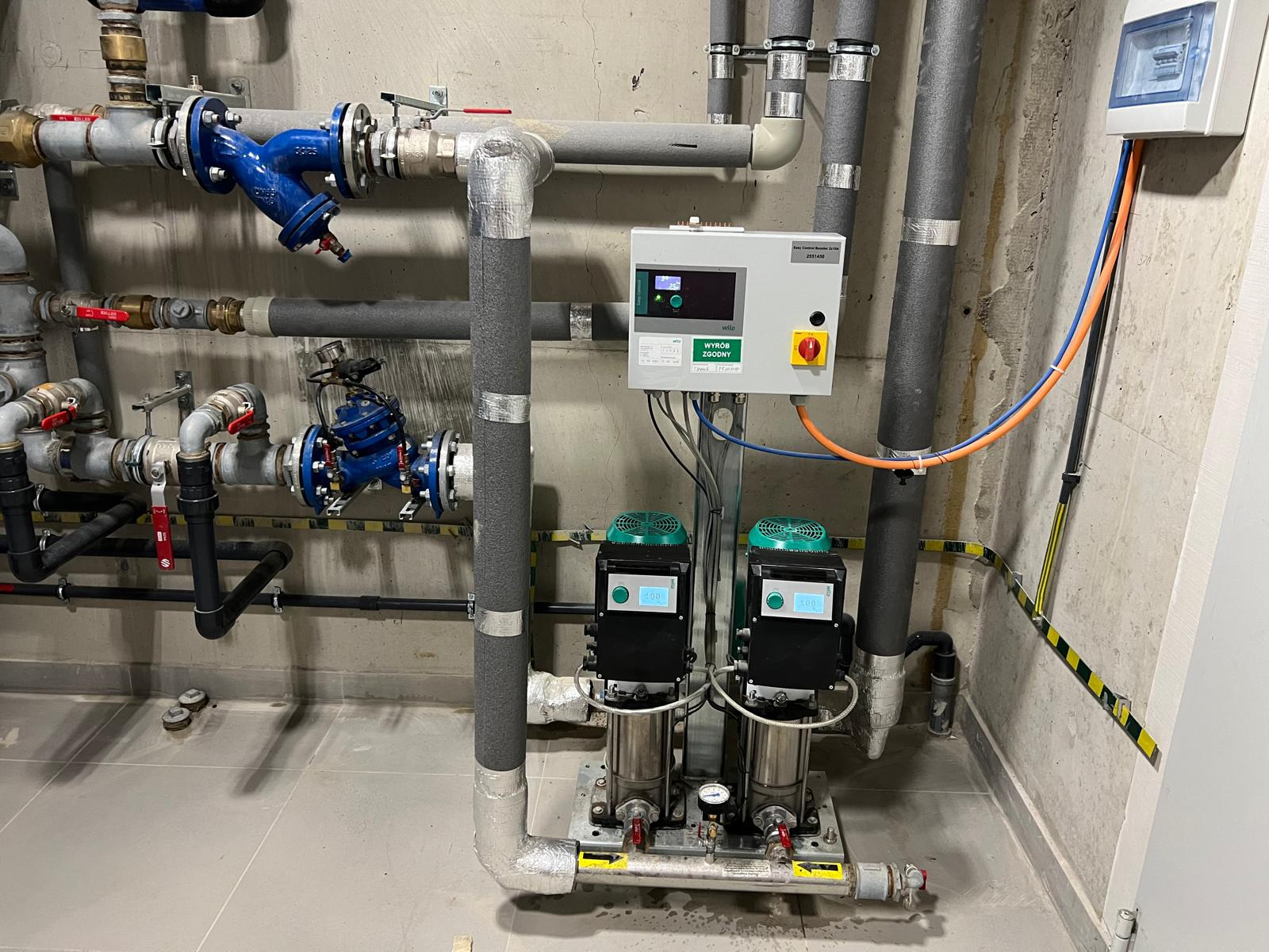 Złożona instalacja hydrauliczna z rurami, zaworami i pompami w pomieszczeniu technicznym. Widoczne izolacje termiczne i panel sterowania. Szara kolorystyka betonu i stali.