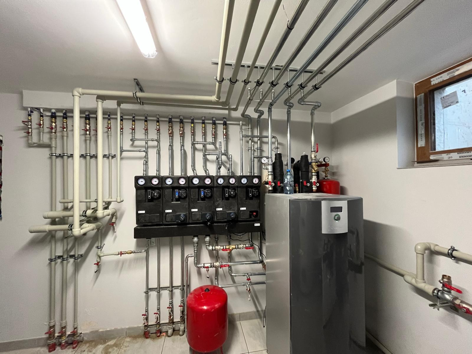 Złożona instalacja grzewcza w kotłowni: liczne rury, zawory, czarne rozdzielacze z manometrami, czerwony zbiornik wyrównawczy i szary piec. Widok na ścianę z oknem.