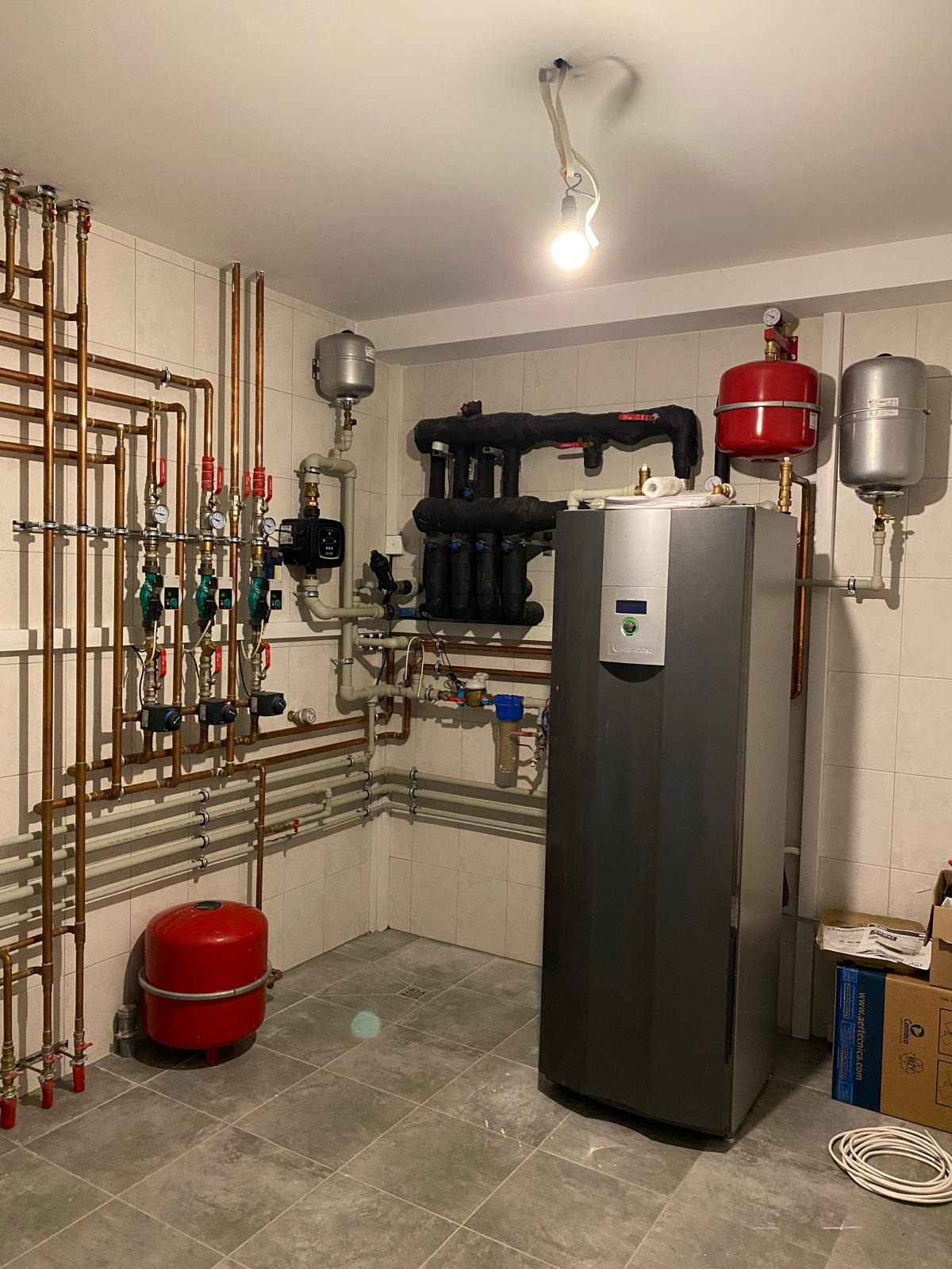 Złożona instalacja CO w kotłowni: nowoczesny piec, liczne miedziane rury, zawory, manometry i zbiorniki wyrównawcze. Białe płytki na ścianach i szara podłoga.