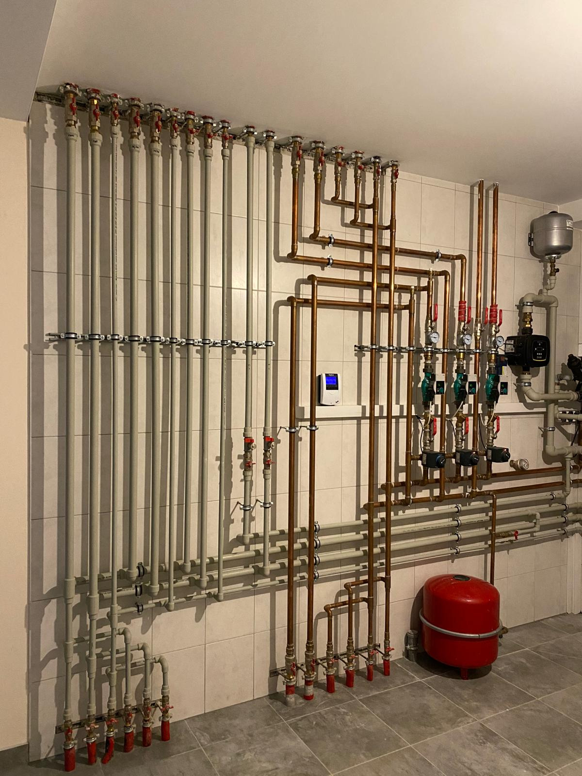 Złożona instalacja grzewcza z rur miedzianych i szarych, zaworów oraz czerwonego zbiornika wyrównawczego na tle białych płytek. Widoczny manometr i sterownik.