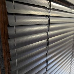 Patryk Kozak Rollkos - Szczegółowe ujęcie perforowanych, szarych żaluzji aluminiowych z widocznym mechanizmem sznurkowym, zamontowanych w oknie z drewnianą ramą.