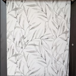 Patryk Kozak Rollkos - Roleta dzień noc z motywem liści na białym tle, zamontowana w czarnej ramie okiennej. Widoczny mechanizm łańcuszkowy po prawej stronie.