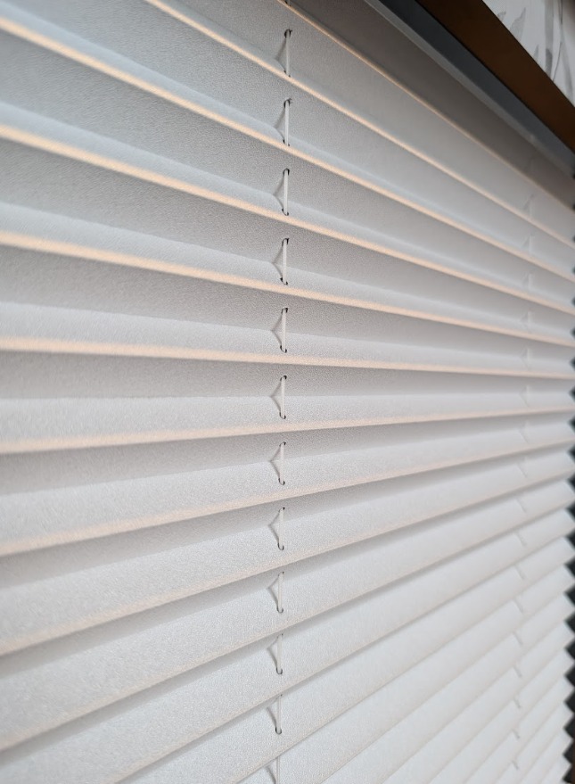 Detal białych plis okiennych z delikatną fakturą, widoczne sznurki i równoległe fałdy, oświetlone miękkim światłem, z fragmentem ramy okiennej w tle.