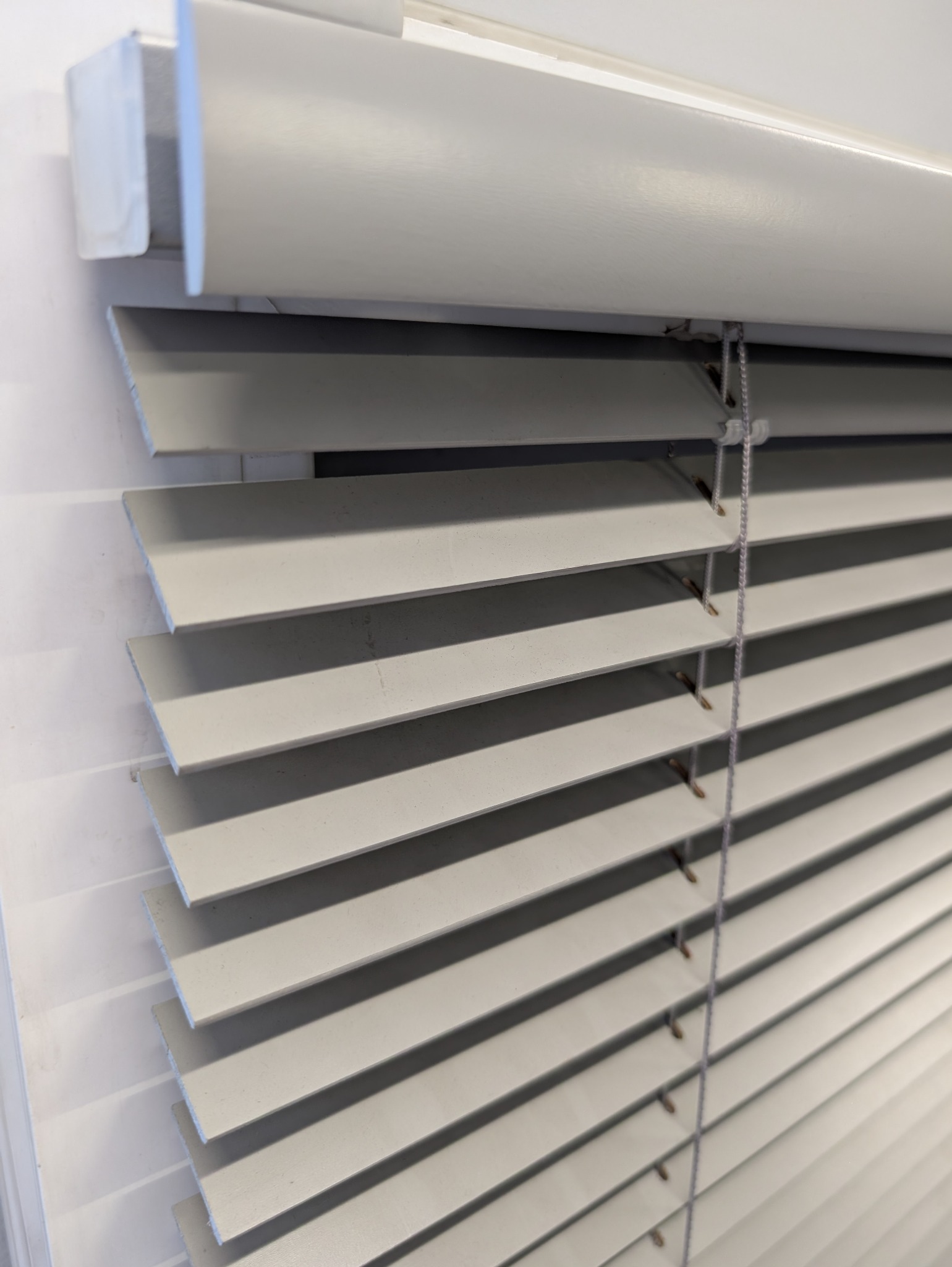 Jasnoszare żaluzje aluminiowe z widocznym mechanizmem sznurkowym, zamontowane w oknie. Delikatne światło podkreśla strukturę lameli.