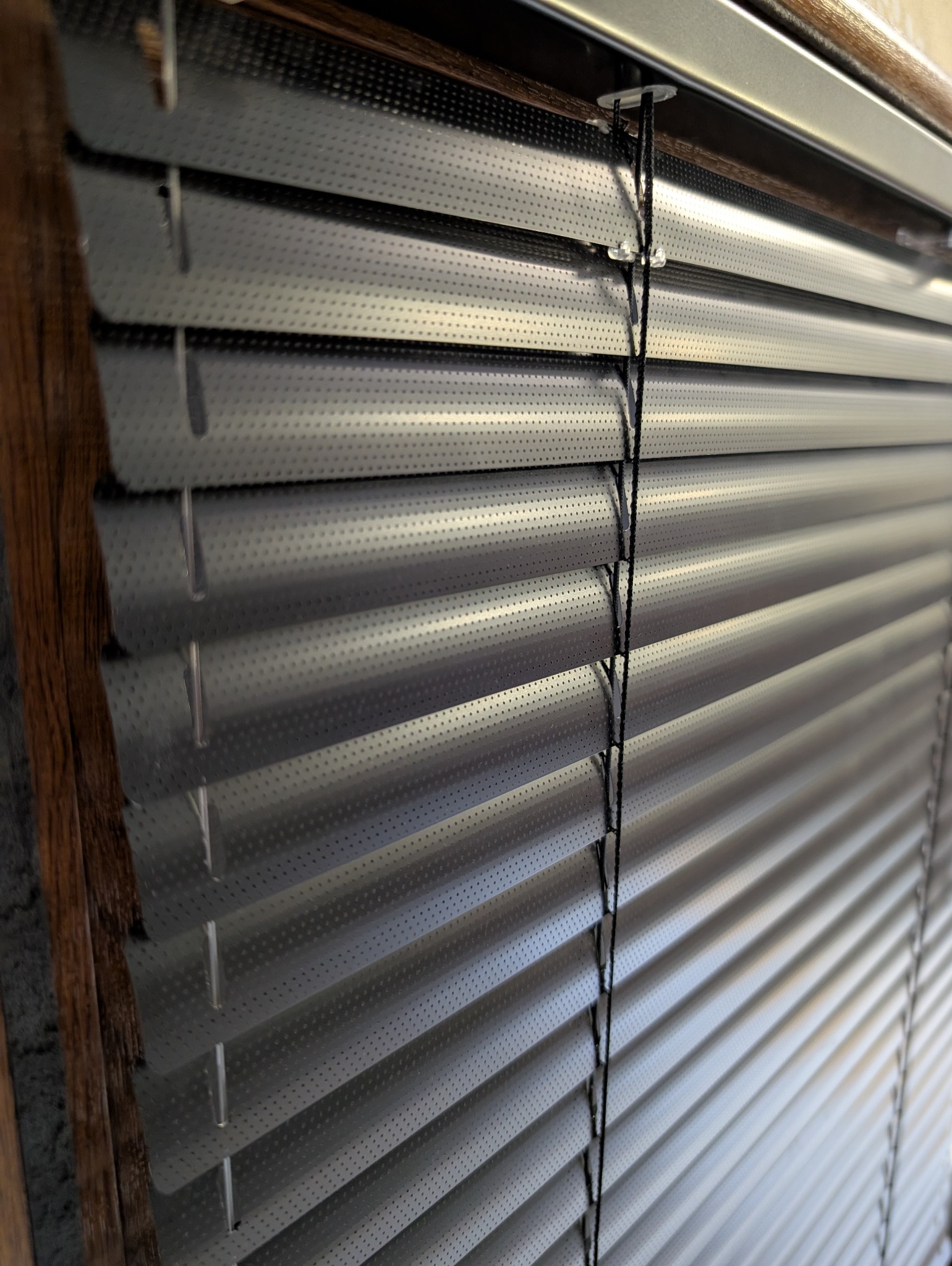 Szczegółowe ujęcie perforowanych, szarych żaluzji aluminiowych z widocznym mechanizmem sznurkowym, zamontowanych w oknie z drewnianą ramą.
