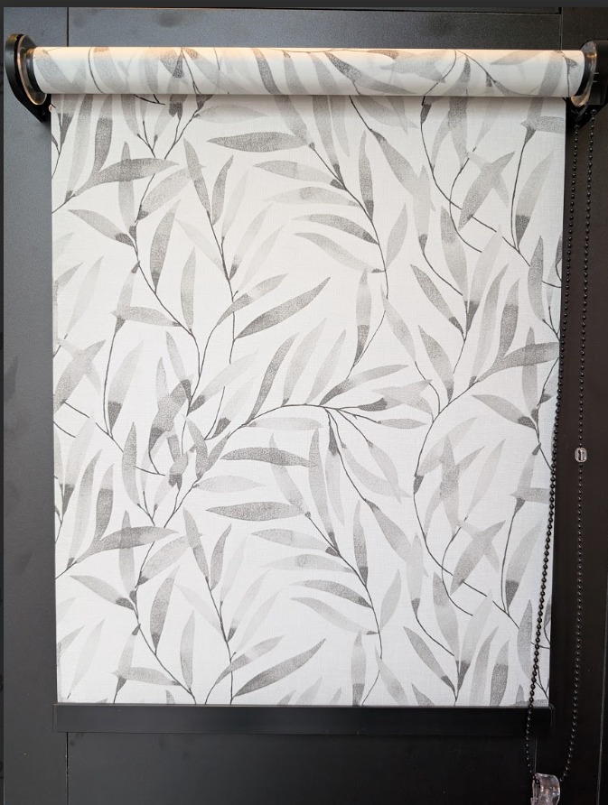 Roleta dzień noc z motywem liści na białym tle, zamontowana w czarnej ramie okiennej. Widoczny mechanizm łańcuszkowy po prawej stronie.