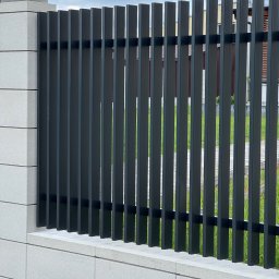 Rafał Ogrodnik OG-MET - Nowoczesne, grafitowe ogrodzenie metalowe z pionowymi sztachetami, osadzone na niskim murku z szarej kostki. Widok na zielony trawnik i fragment budynku w tle.