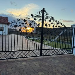 Rafał Ogrodnik OG-MET - Otwarta, kuta brama wjazdowa z motywem roślinnym na tle zachodzącego słońca. Widoczny fragment domu i kostka brukowa na podjeździe. Dekoracyjne wykończenie.