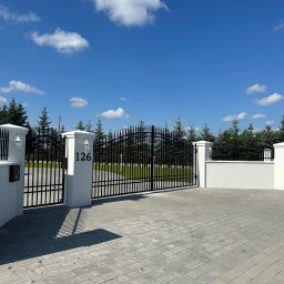 Rafał Ogrodnik OG-MET - Czarna brama wjazdowa z numerem 126 na białym słupku, otoczona białymi murkami i słupkami z lampami. Kostka brukowa na podjeździe, w tle drzewa i błękitne niebo.