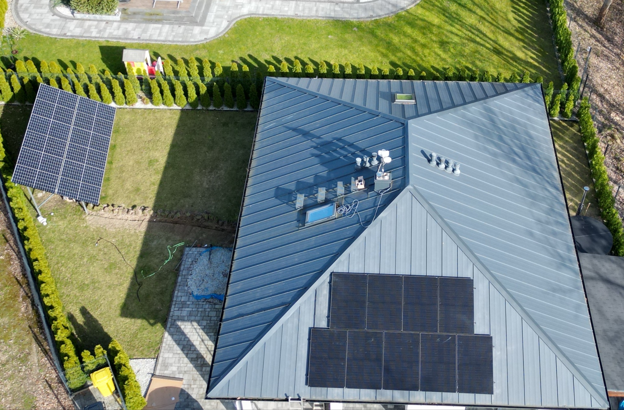 Widok z góry na dom z panelami słonecznymi na dachu i w ogrodzie. Instalacja fotowoltaiczna na metalowym dachu, obok trawnik i ozdobne krzewy.