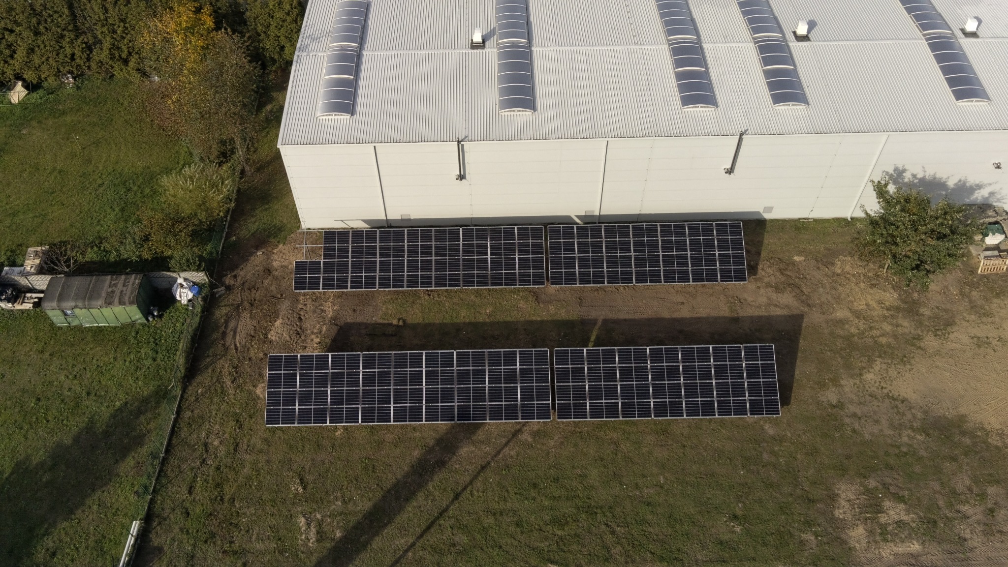 Widok z góry na instalację fotowoltaiczną na dachu magazynu z panelami ułożonymi w dwóch rzędach, otoczoną zielenią i drzewami. Dach z świetlikami.