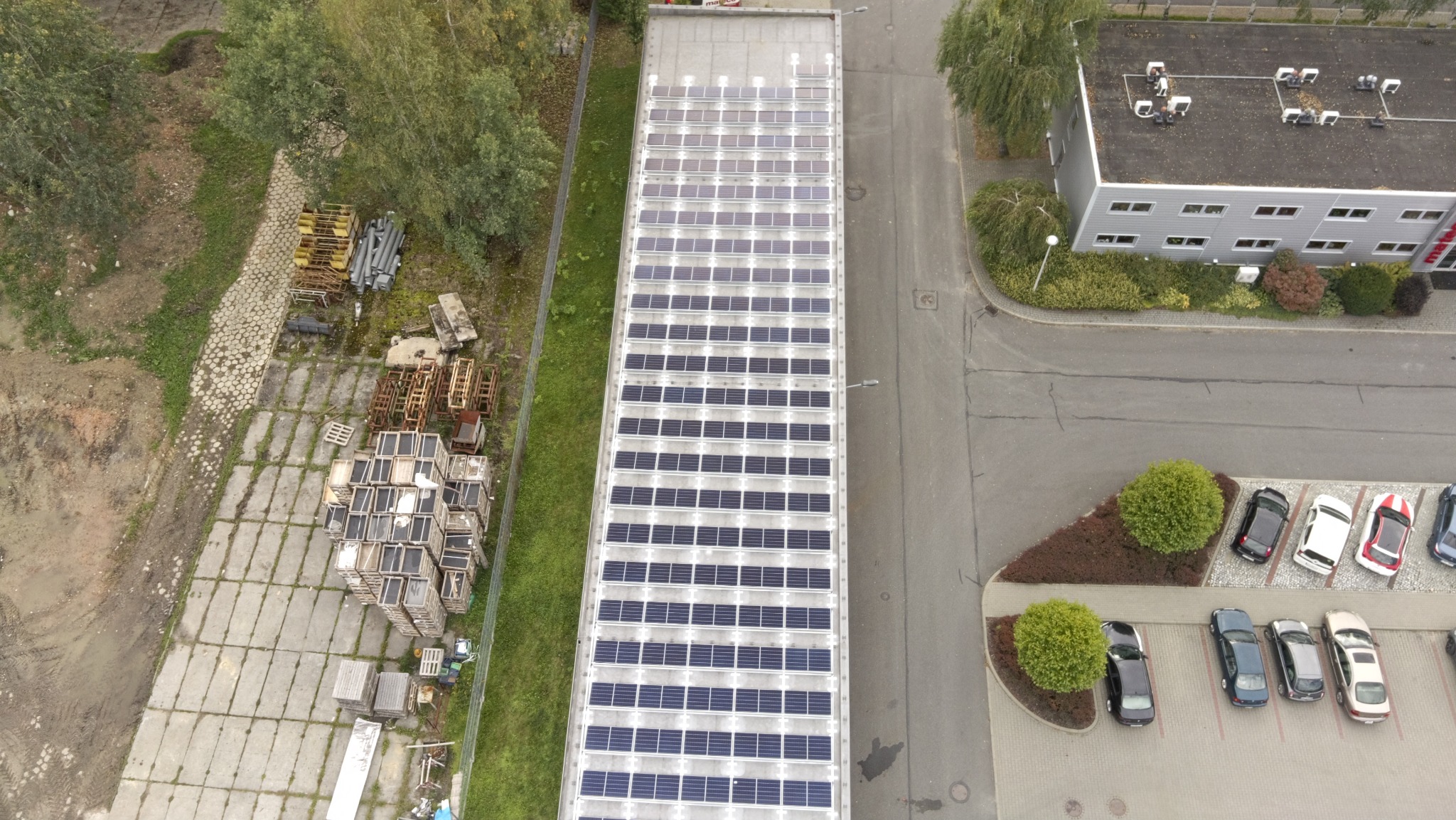 Widok z góry na dach budynku z panelami słonecznymi, obok parking i zieleń. Na drugim planie budynek biurowy i drzewa. Ujęcie z drona, pochmurny dzień.