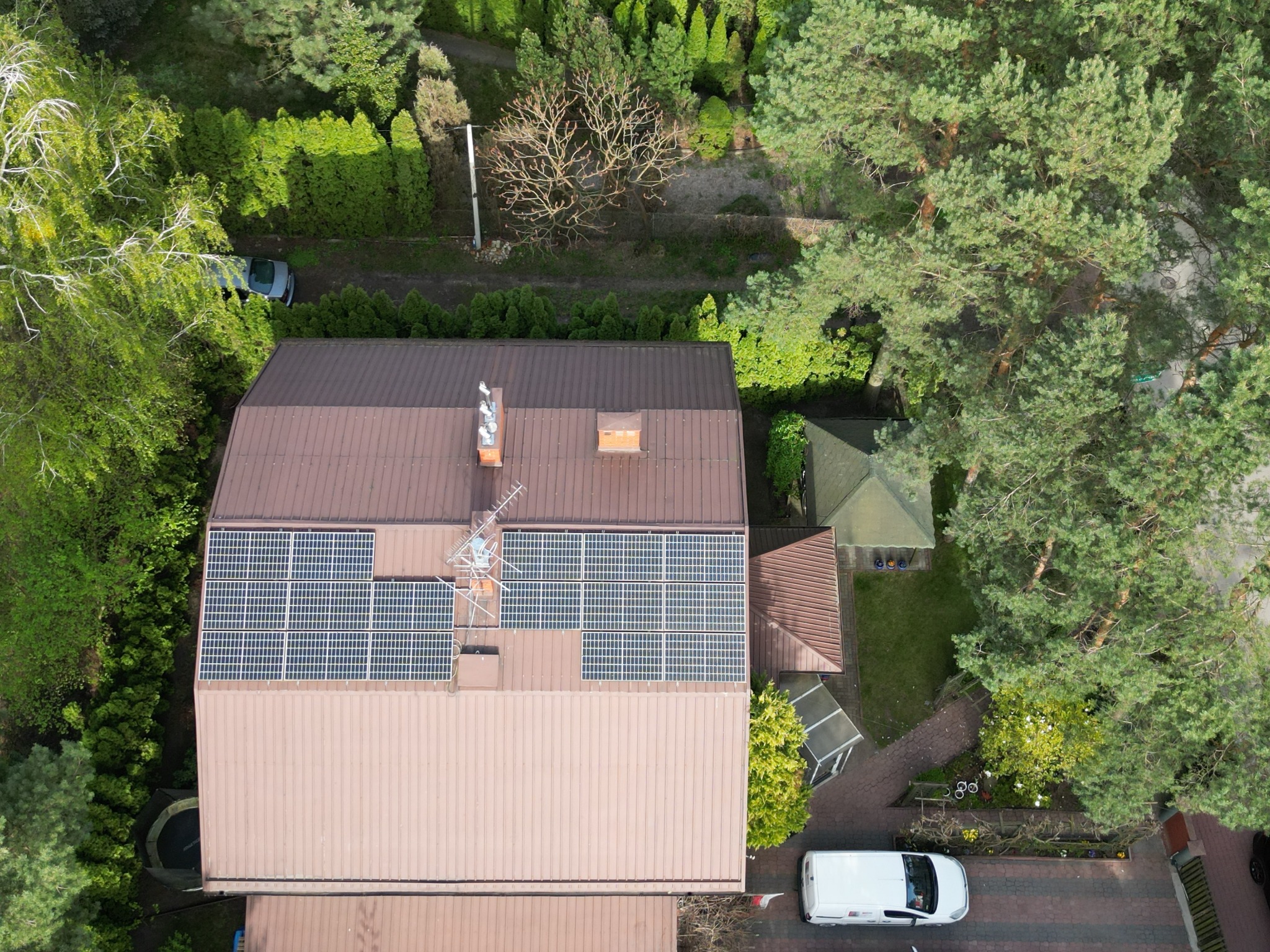 Widok z drona na dom z brązowym dachem i panelami fotowoltaicznymi. Wokół domu bujna zieleń, drzewa i zaparkowane samochody. Ujęcie z góry.