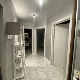 KOSTAGO Kostiantyn Kostenko - Korytarz po remoncie: białe marmurowe płytki podłogowe, szare ściany z efektem dekoracyjnym, białe drzwi i sufit napinany z oświetleniem punktowym.