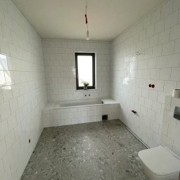 KOSTAGO Kostiantyn Kostenko - Jasna łazienka po remoncie: białe, prostokątne płytki na ścianach, szara podłoga z wzorem lastryko, wanna obudowana płytkami, okno z rośliną, minimalistyczna toaleta.