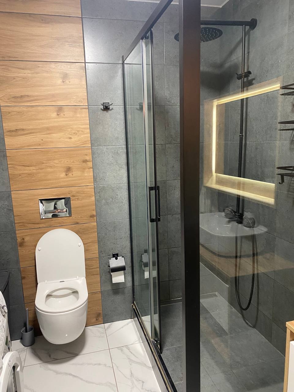 Nowoczesna łazienka po remoncie: szare płytki, drewniane akcenty, kabina prysznicowa z czarną ramą i podświetlane lustro nad umywalką. Biała toaleta i marmurowe płytki podłogowe.