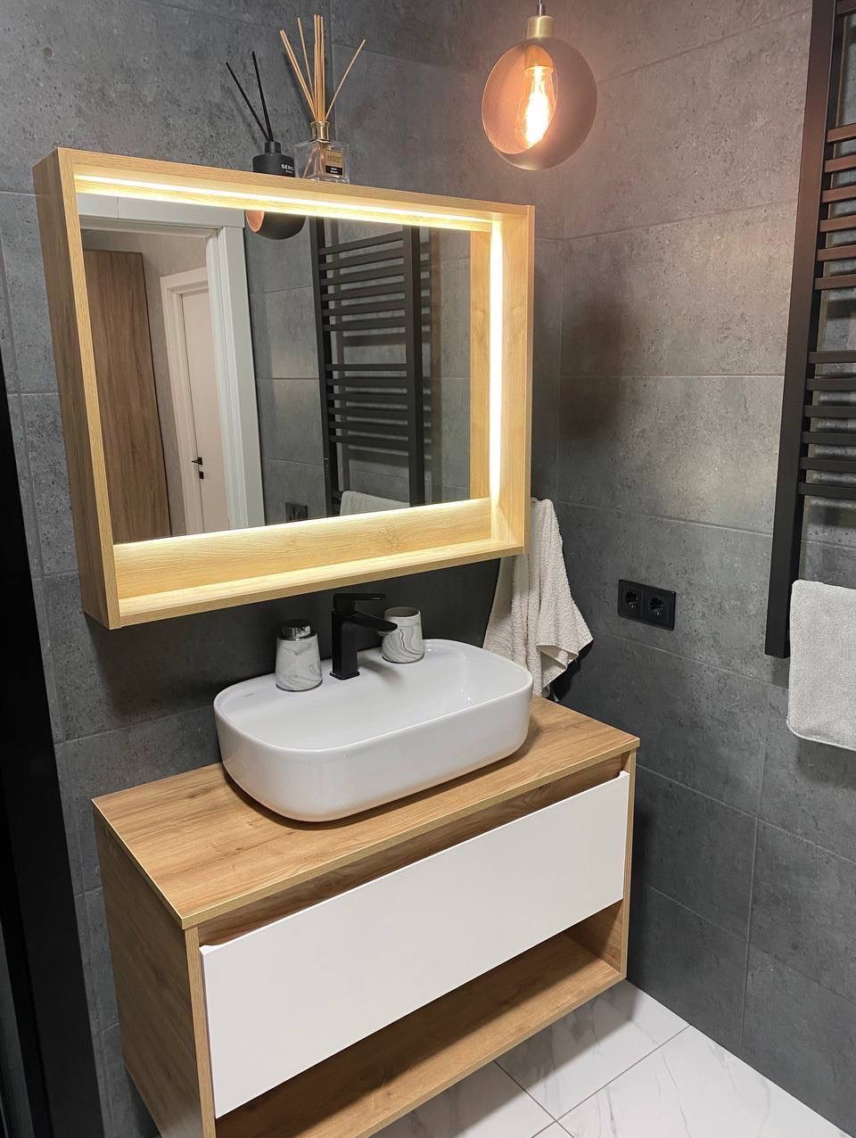 Nowoczesna łazienka z podświetlanym lustrem, umywalką nablatową i szarą płytką. Minimalistyczny design z drewnianymi akcentami i dekoracyjnym oświetleniem.