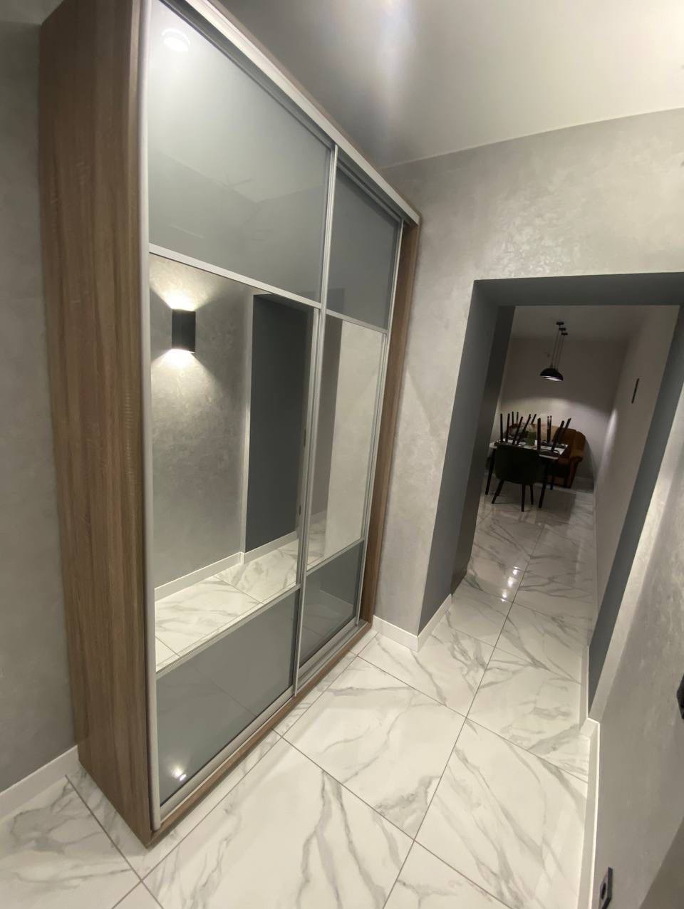 Szafa przesuwna z lustrami w przedpokoju, oświetlenie punktowe, marmurowe płytki podłogowe. Widok na jadalnię w głębi. Wykończenie wnętrz w szarościach i bieli.