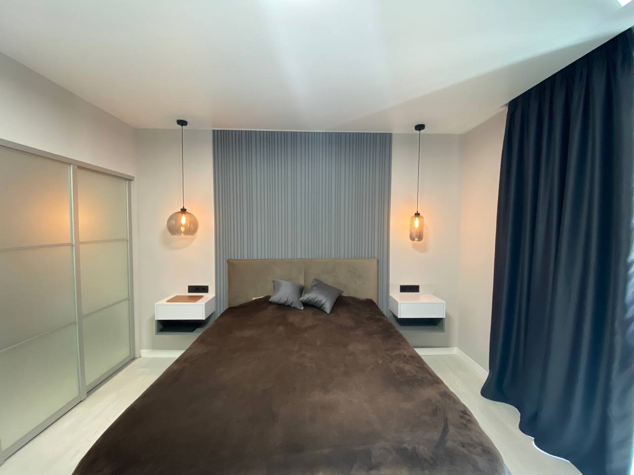 Nowoczesna sypialnia z łóżkiem, szarą ścianą z panelami, wiszącymi lampami i przesuwną szafą. Minimalistyczny design z akcentami kolorystycznymi.