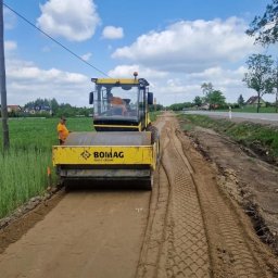Modum Sp. z o. o. - Żółty walec drogowy BOMAG zagęszcza piaszczysty grunt pod budowę nowej drogi, obok istniejącej asfaltowej jezdni. Pracownik w pomarańczowej koszulce nadzoruje prace.