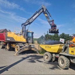 Modum Sp. z o. o. - Żółta koparka Volvo EW160B Modum ładuje masę bitumiczną do miniwywrotki Wacker Neuson, obsługiwanej przez pracownika. W tle czerwona ciężarówka i domy.