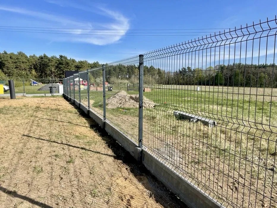 Nowoczesne ogrodzenie panelowe z podmurówką betonową na tle zieleni i błękitnego nieba. Widoczne słupki i siatka ogrodzeniowa. Ujęcie z bliska, podkreślające detale.