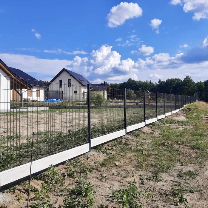 Nowoczesne ogrodzenie panelowe z podmurówką betonową, widok z zewnątrz posesji, na tle domów jednorodzinnych i błękitnego nieba z chmurami.