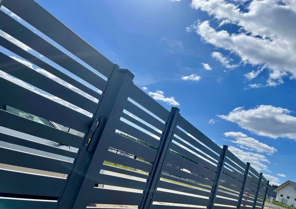 Nowoczesne, poziome przęsła ogrodzeniowe w kolorze grafitowym na tle błękitnego nieba z chmurami. Widoczny zamek w furtce i fragment domu w oddali.