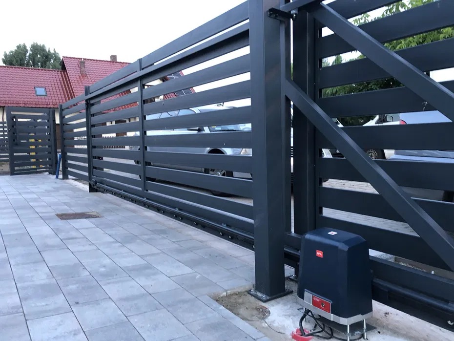 Nowoczesna, przesuwna brama wjazdowa z automatyką, szara, poziome panele, widok z zewnątrz na posesję z kostką brukową i domem w tle. Brama zamontowana.