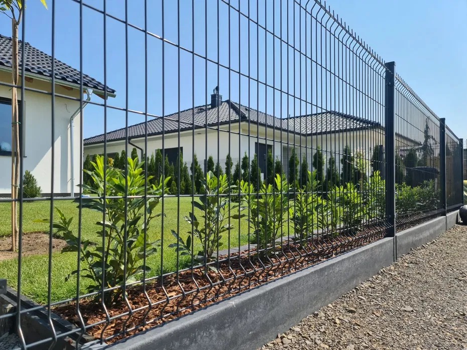 Nowoczesne ogrodzenie panelowe w kolorze grafitowym, z podmurówką i nasadzeniami z roślin, na tle zadbanego trawnika i domu jednorodzinnego w słoneczny dzień.