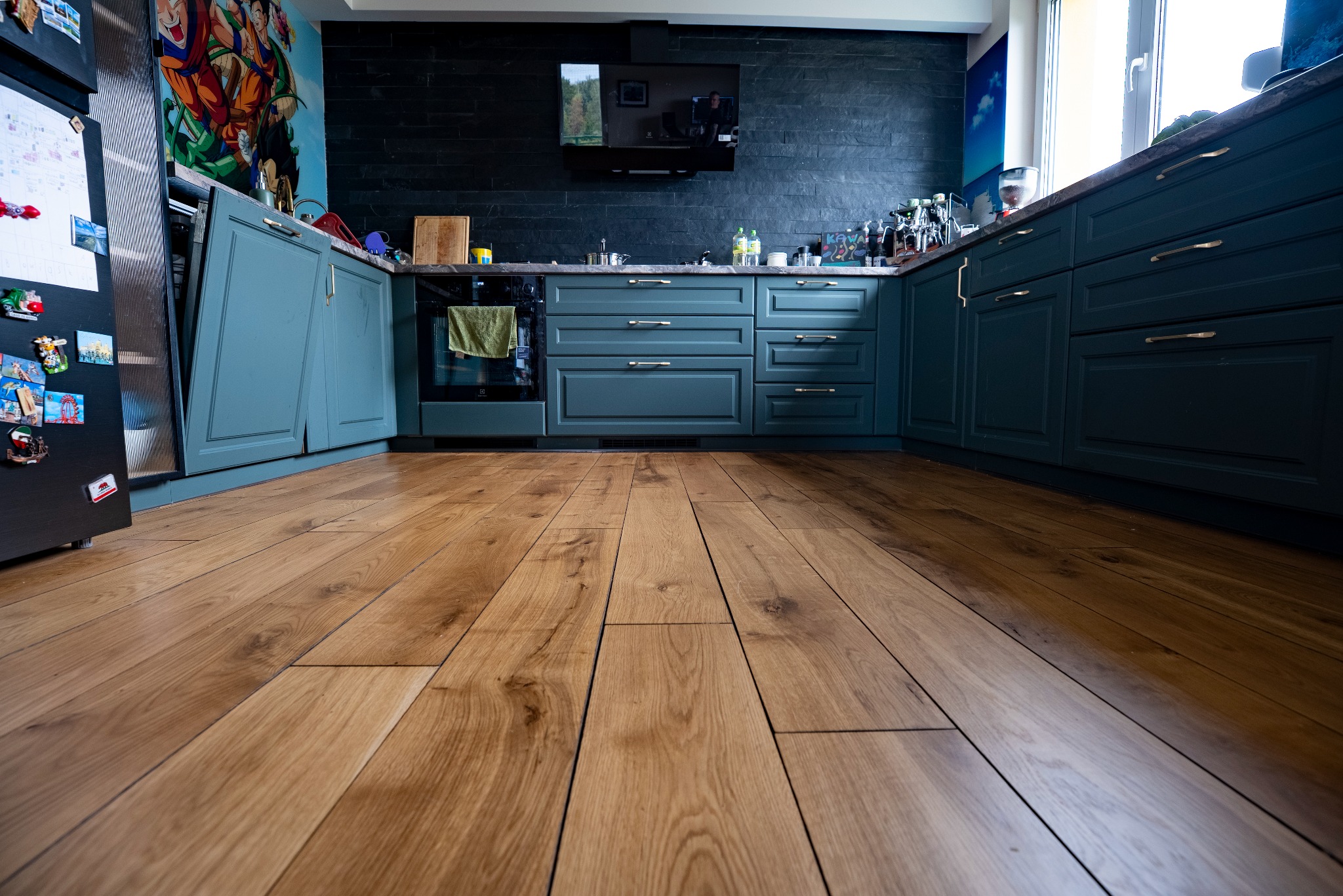 Jasne, drewniane panele podłogowe w kuchni z ciemnymi, eleganckimi szafkami i czarną ścianą z telewizorem. Uwagę przykuwa oryginalna grafika na ścianie obok lodówki.