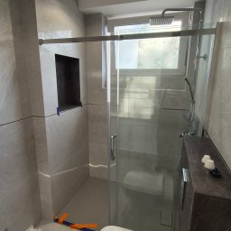 ZAKŁAD REMONTOWO-BUDOWLANY REMNAL PIOTR NALEWAJKA - Nowoczesna łazienka z kabiną prysznicową typu walk-in, szarą glazurą i podtynkową armaturą. Widoczny fragment toalety i oświetlenie sufitowe.