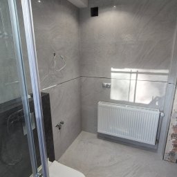 ZAKŁAD REMONTOWO-BUDOWLANY REMNAL PIOTR NALEWAJKA - Jasna łazienka z szarymi płytkami imitującymi kamień, kabiną prysznicową ze szkła i białym grzejnikiem. Widoczna toaleta podwieszana i minimalistyczne oświetlenie sufitowe.