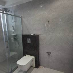 ZAKŁAD REMONTOWO-BUDOWLANY REMNAL PIOTR NALEWAJKA - Nowoczesna łazienka z kabiną prysznicową i podwieszaną toaletą. Szare płytki imitujące kamień, chromowane detale, minimalistyczny design. Widok na grzejnik.