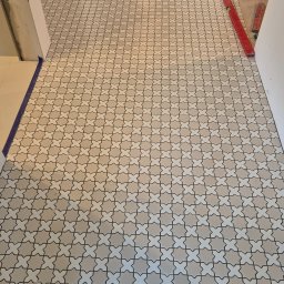 ZAKŁAD REMONTOWO-BUDOWLANY REMNAL PIOTR NALEWAJKA - Ułożona, wzorzysta podłoga z płytek w odcieniach beżu i bieli, widok z perspektywy wejścia do pomieszczenia. Czerwona poziomica leży przy ścianie.