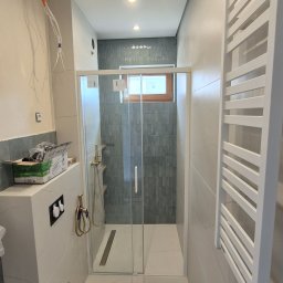 ZAKŁAD REMONTOWO-BUDOWLANY REMNAL PIOTR NALEWAJKA - Nowoczesna łazienka z kabiną prysznicową typu walk-in, wykończona płytkami w odcieniach szarości i bieli. Widoczne elementy armatury w kolorze złotym i grzejnik drabinkowy.