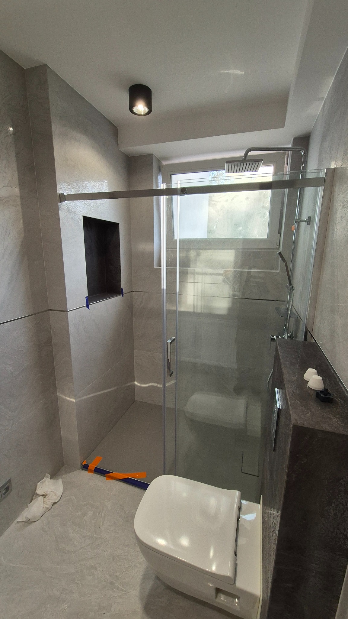 Nowoczesna łazienka z kabiną prysznicową typu walk-in, szarą glazurą i podtynkową armaturą. Widoczny fragment toalety i oświetlenie sufitowe.