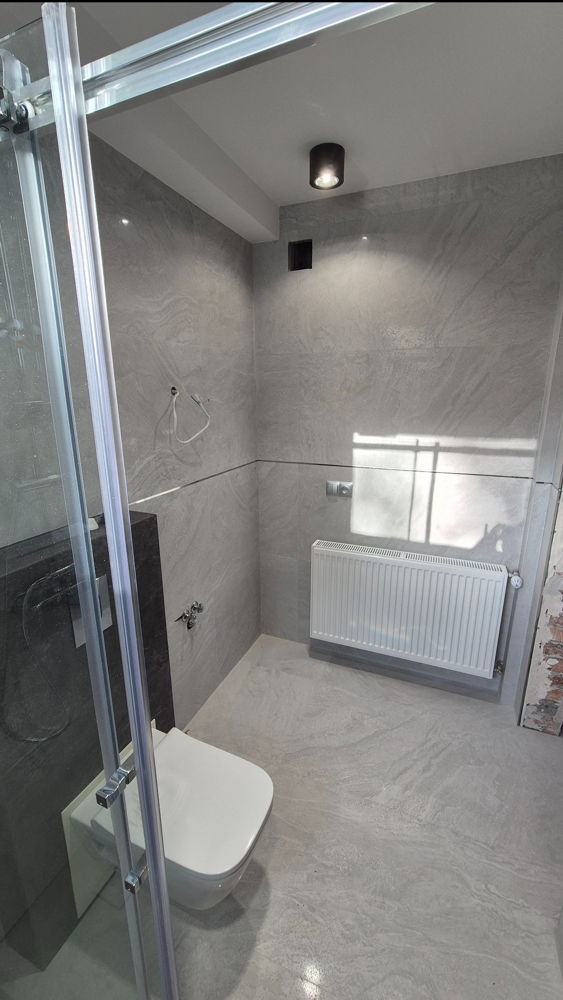 Jasna łazienka z szarymi płytkami imitującymi kamień, kabiną prysznicową ze szkła i białym grzejnikiem. Widoczna toaleta podwieszana i minimalistyczne oświetlenie sufitowe.