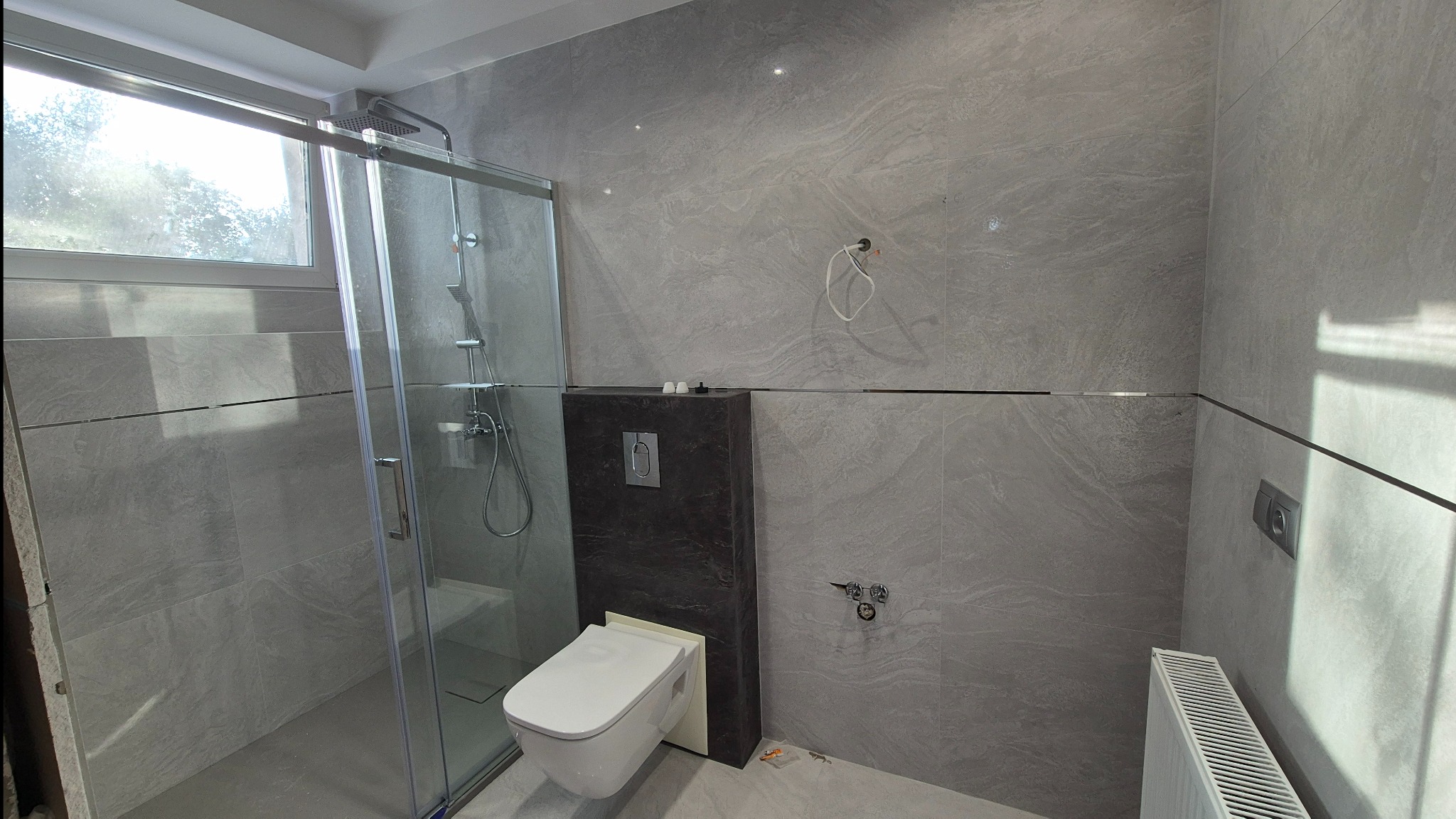 Nowoczesna łazienka z kabiną prysznicową i podwieszaną toaletą. Szare płytki imitujące kamień, chromowane detale, minimalistyczny design. Widok na grzejnik.