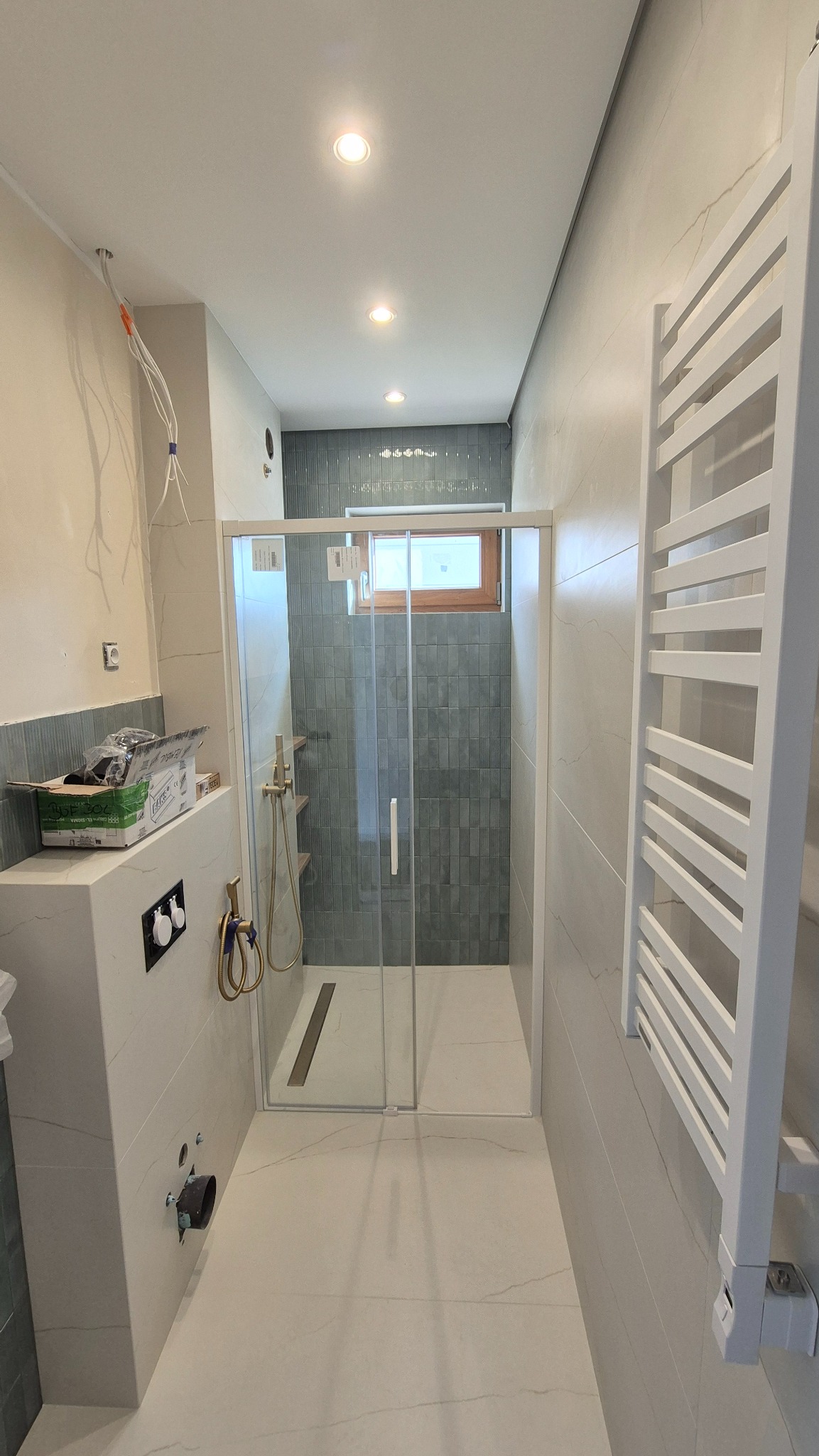 Nowoczesna łazienka z kabiną prysznicową typu walk-in, wykończona płytkami w odcieniach szarości i bieli. Widoczne elementy armatury w kolorze złotym i grzejnik drabinkowy.