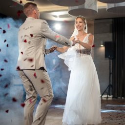 Maja Jurkiewicz Fotografia - Para młoda tańczy w obłoku konfetti w kształcie serc. Panna młoda w tiarze i sukni z koronkową górą, pan młody w beżowym garniturze. W tle zespół muzyczny i dekoracje.