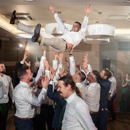 Maja Jurkiewicz Fotografia - Radosny pan młody uniesiony w górę przez gości weselnych podczas zabawy na sali. Widoczne oświetlenie sufitowe i tańczący goście w tle.