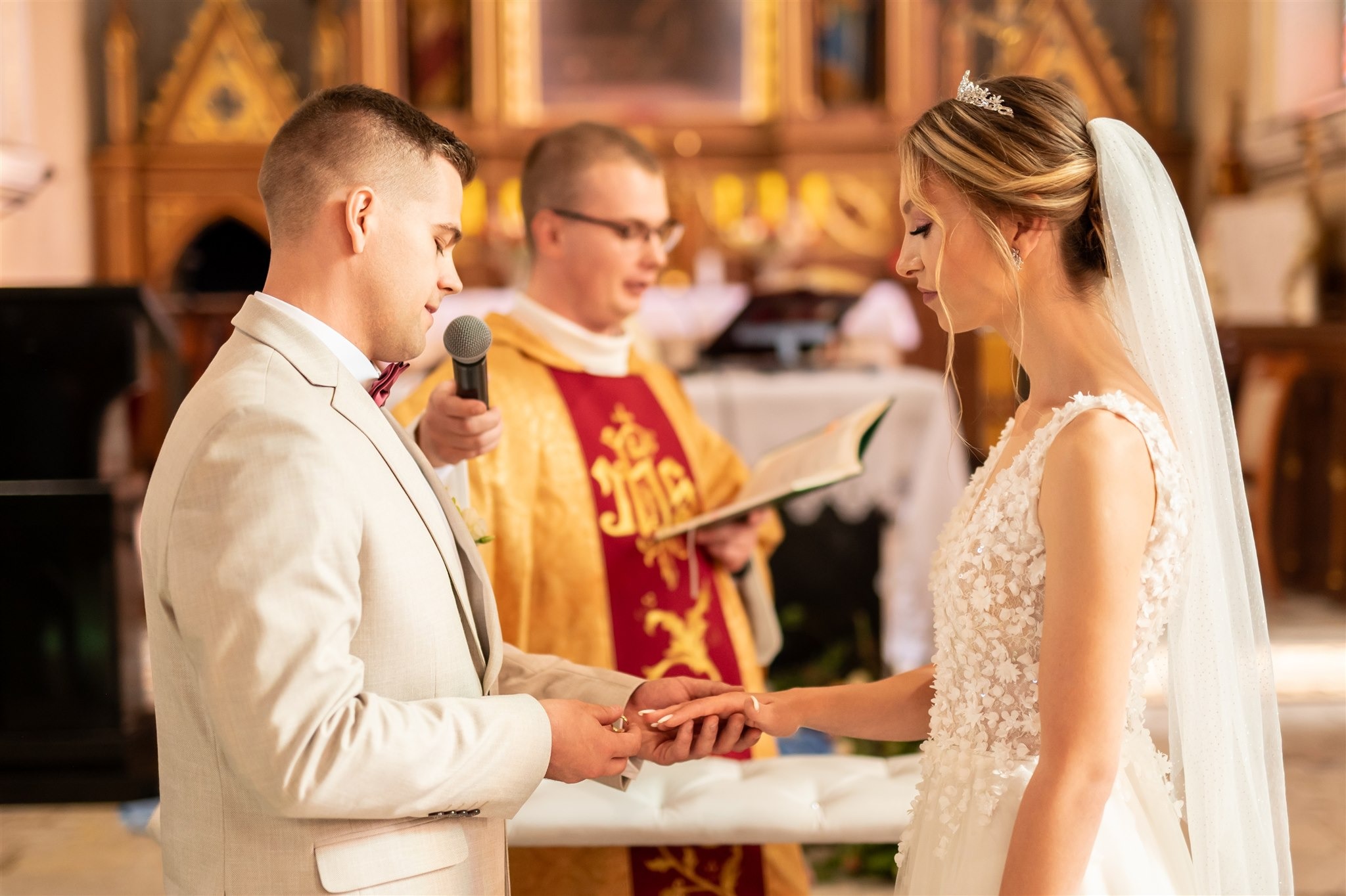 Moment zakładania obrączki podczas ceremonii ślubnej w kościele. Panna młoda w białej sukni, pan młody w beżowym garniturze. W tle ksiądz w szatach liturgicznych.