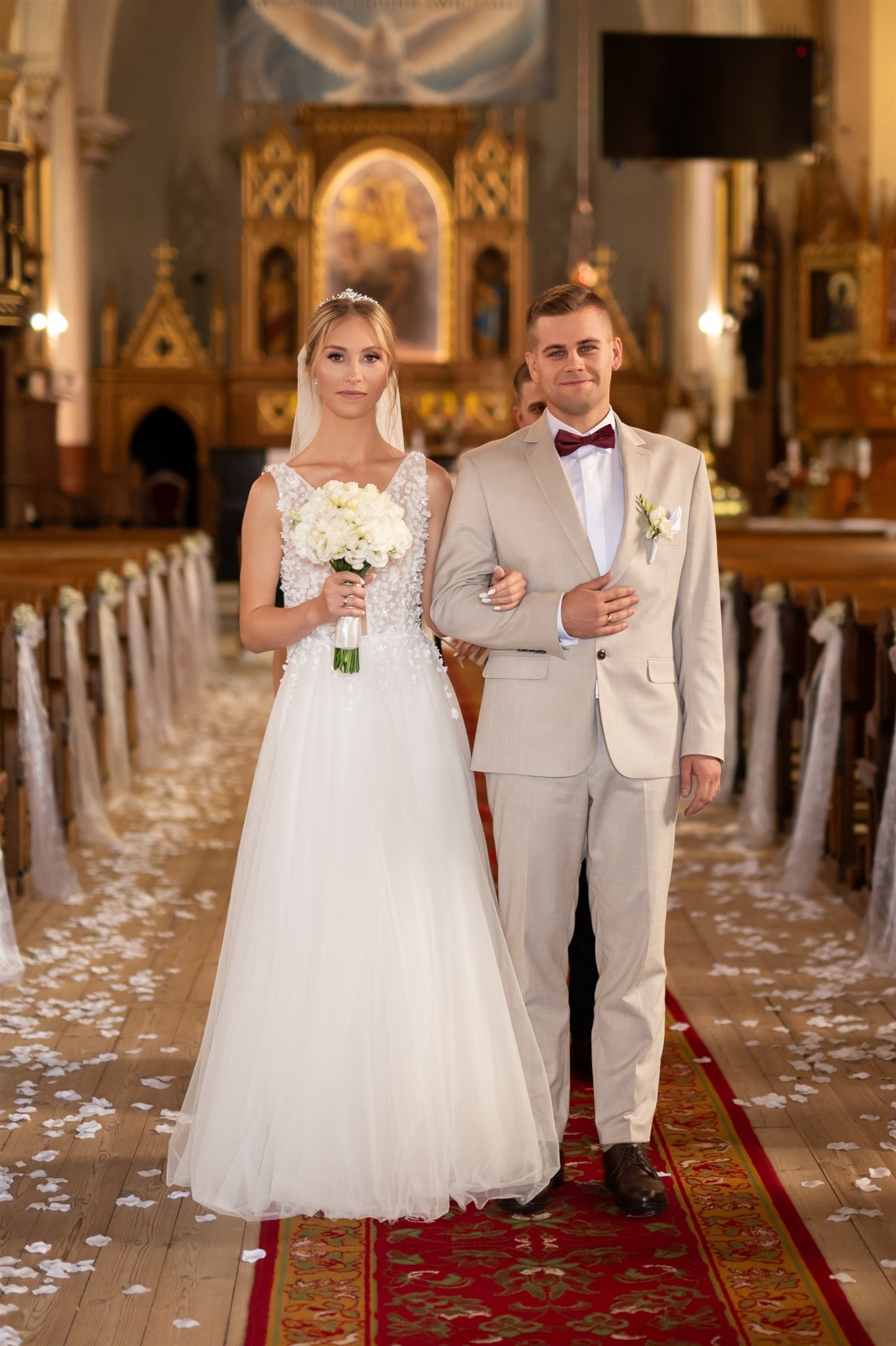 Para młoda w kościele: panna młoda w białej sukni z bukietem, pan młody w beżowym garniturze i bordowej muszce. W tle ołtarz i ławki z dekoracjami.