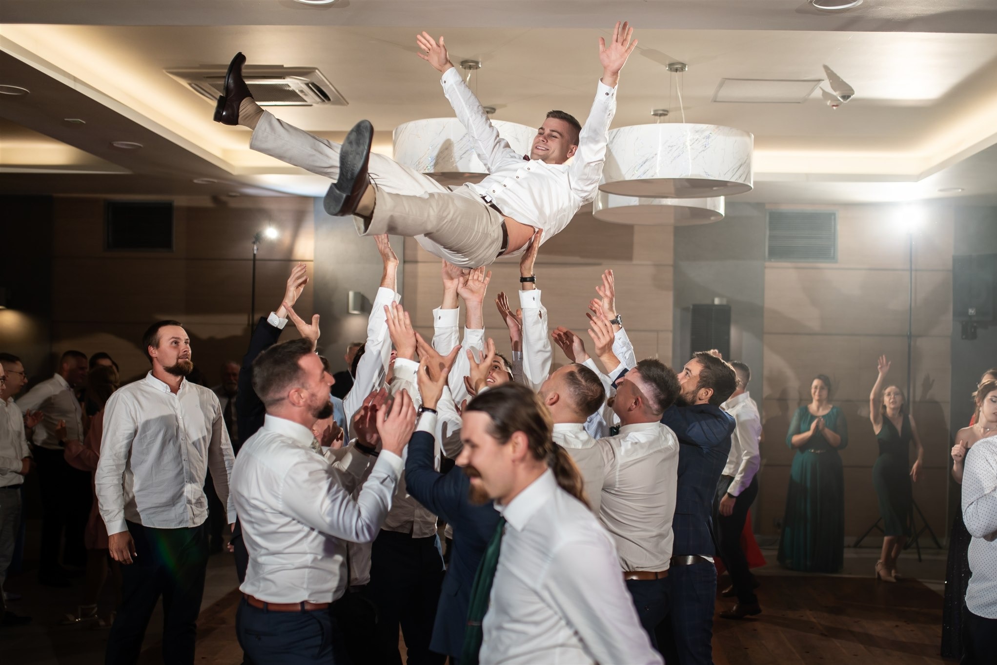 Radosny pan młody uniesiony w górę przez gości weselnych podczas zabawy na sali. Widoczne oświetlenie sufitowe i tańczący goście w tle.