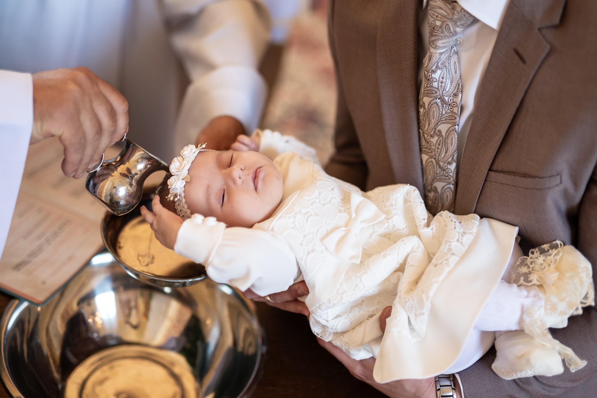 Ujęcie z ceremonii chrztu: śpiące niemowlę w białej sukience, trzymane przez mężczyznę w garniturze, nad nim dłoń kapłana z naczyniem do polewania wodą.
