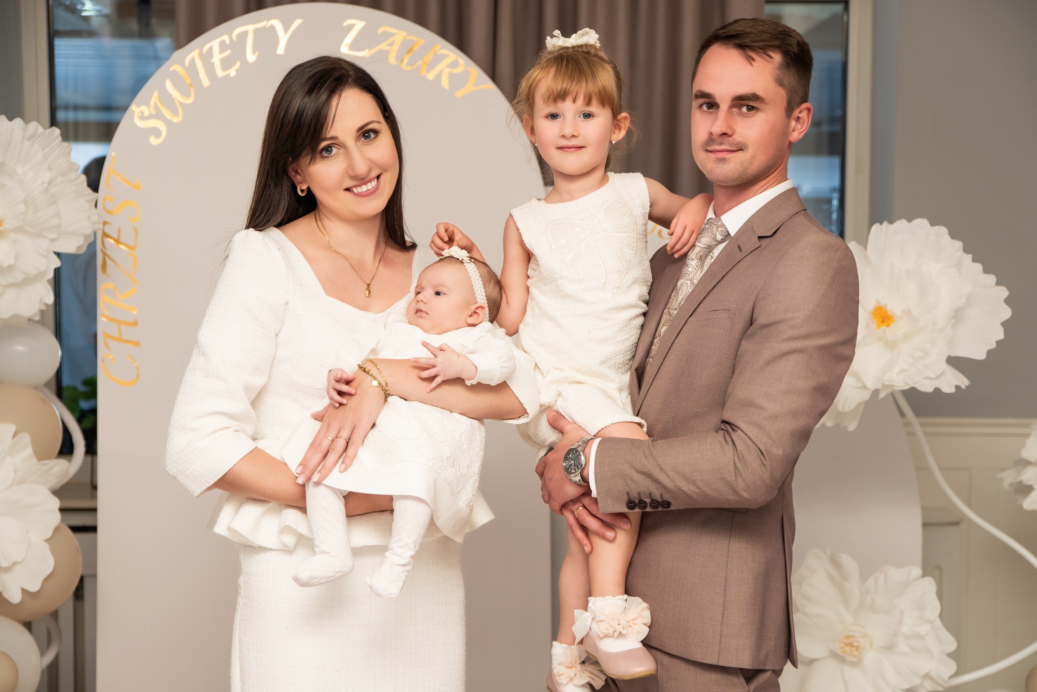 Uśmiechnięta rodzina w eleganckich strojach pozuje na tle dekoracji z napisem 'Święty Chrzest Laury' i papierowych kwiatów. Mama trzyma niemowlę, tata starszą córkę.
